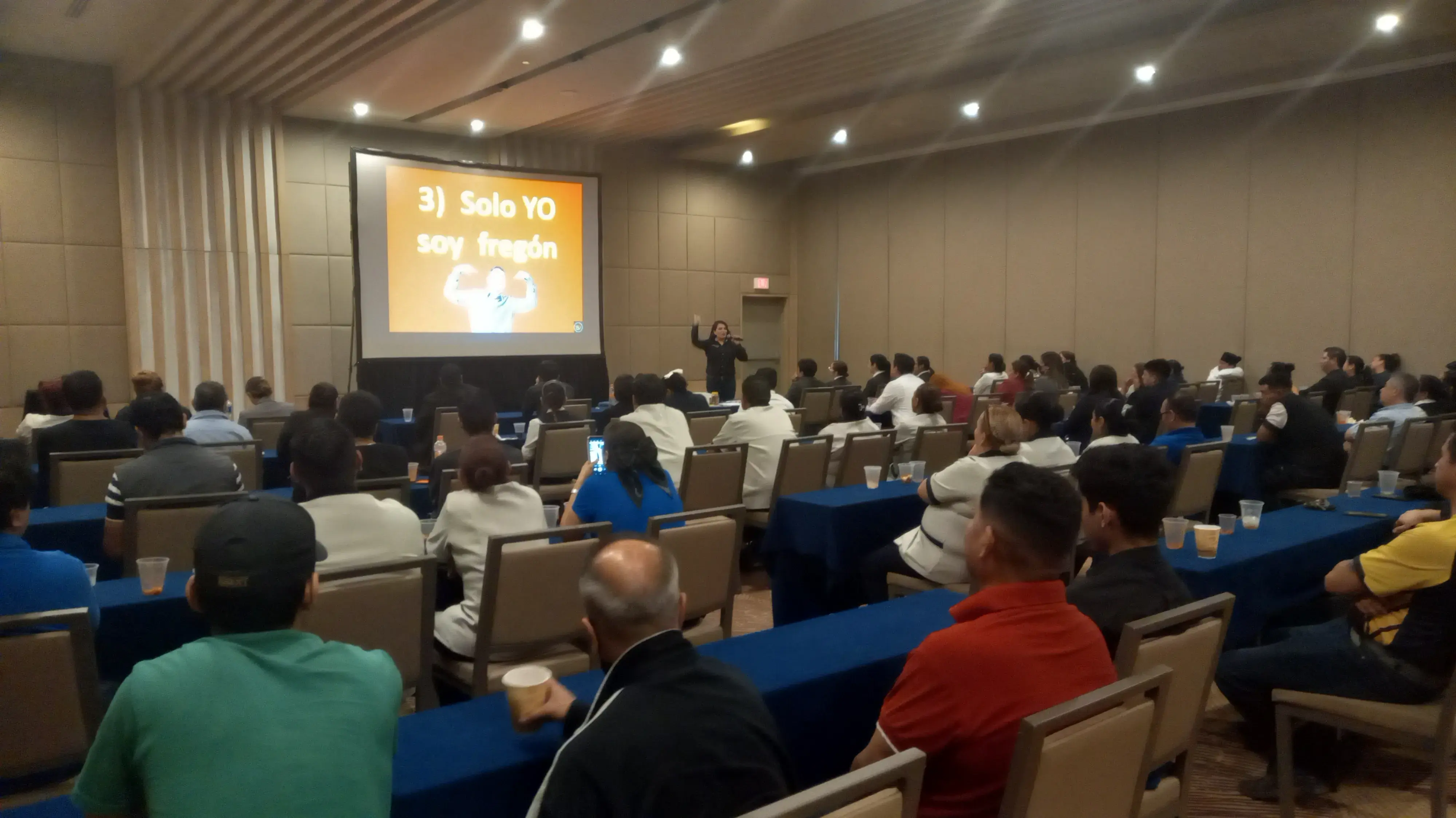 Entrenador empresarial exponiendo ante un auditorio lleno, dentro de un centro de entrenamiento empresarial enfocado en desarrollo profesional y liderazgo. 