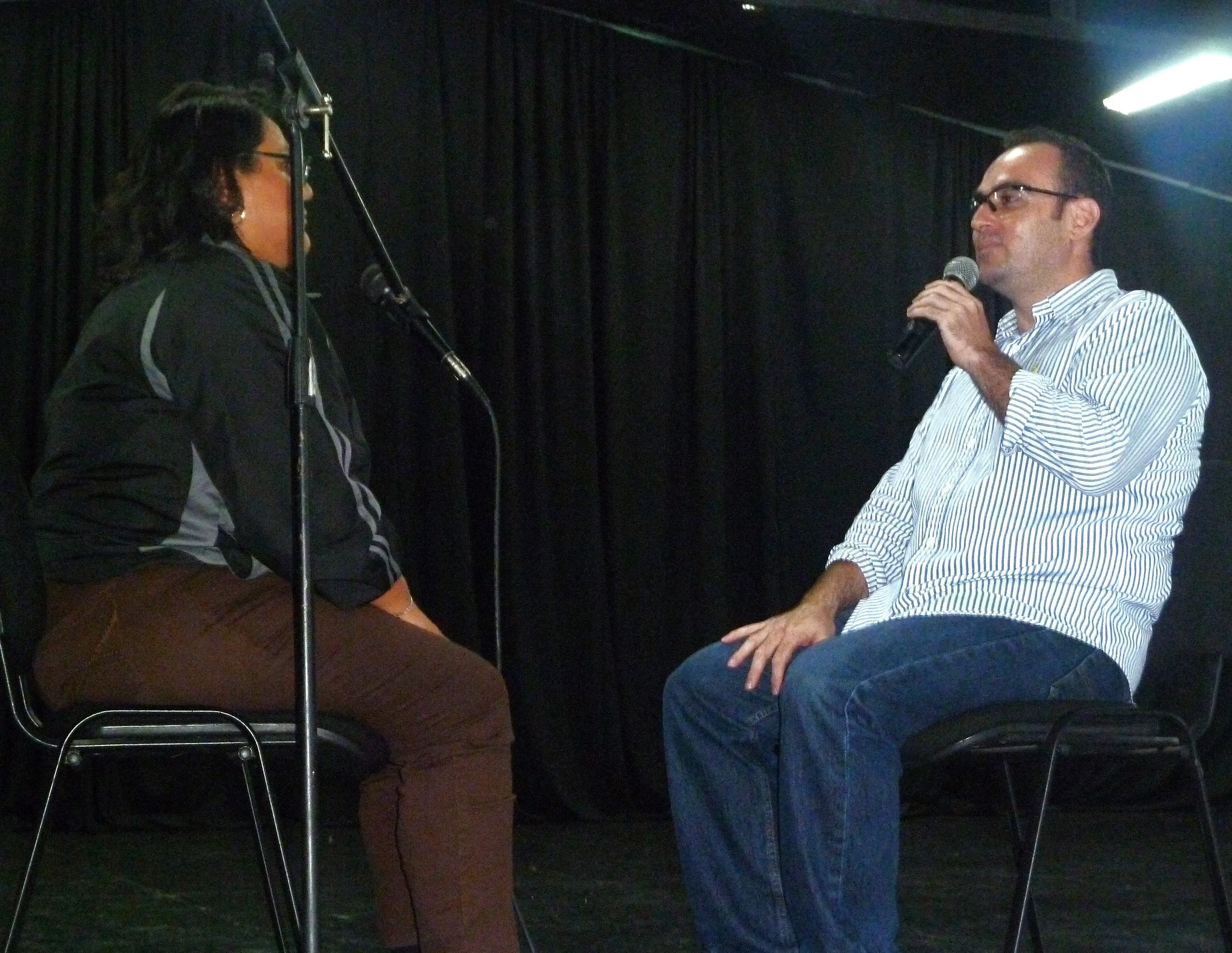 Hombre en escenario capacitando sobre un curso de ventas Fotografía de una mujer sentada y un Hombre sentado arriba de un escenario capacitando sobre un curso de ventas